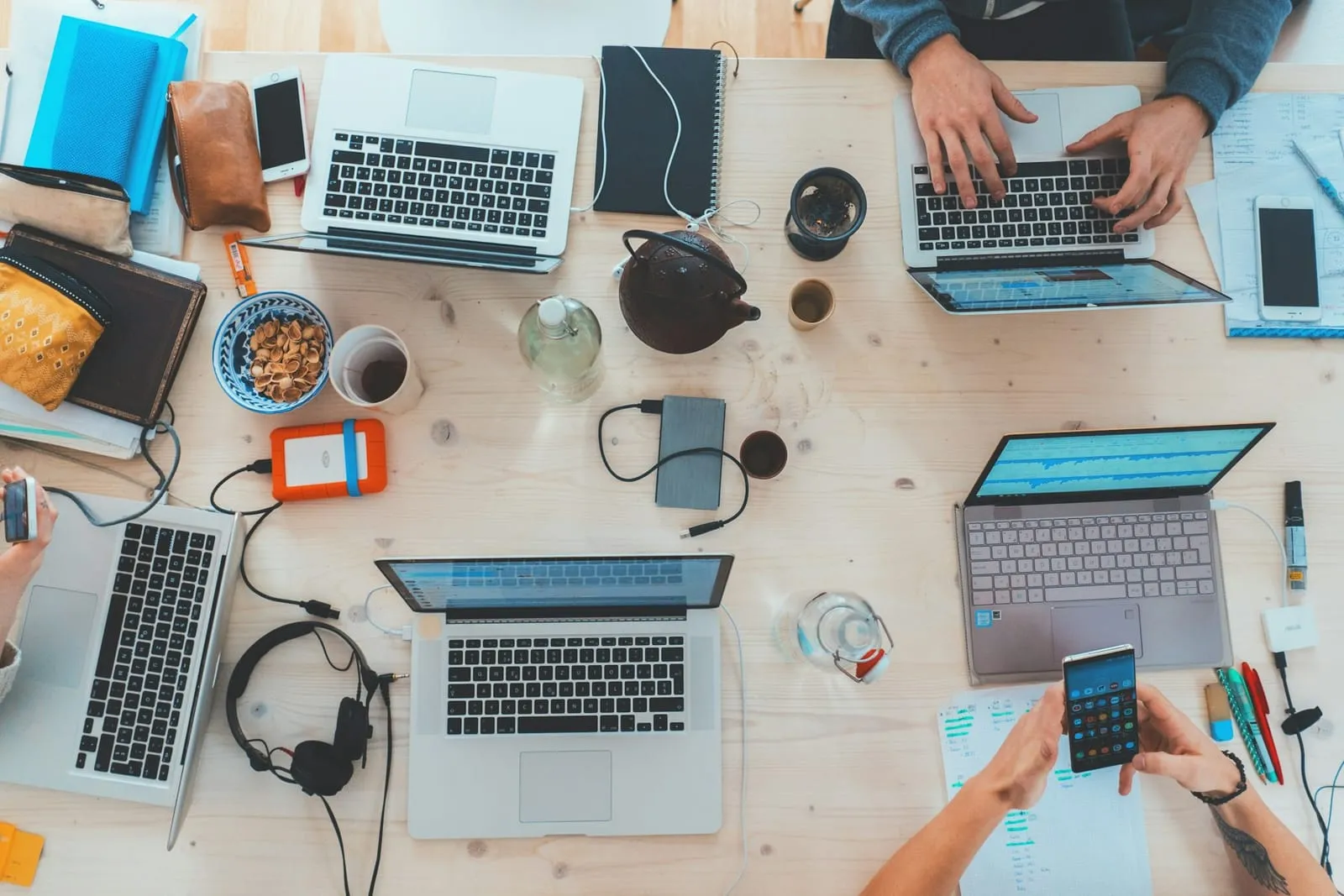 Overhead view of a shared table with several laptops, notebooks, phones and cups of coffee as a team works together