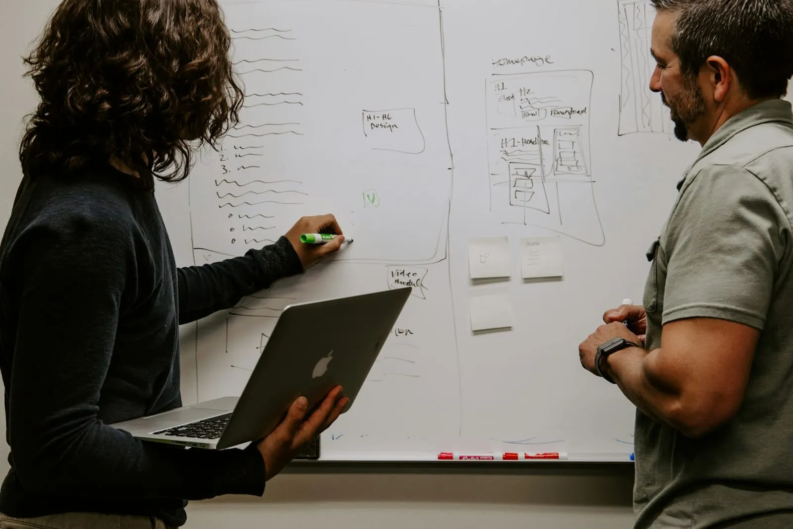 Two colleagues stand at a whiteboard covered in sketched plans, one of them holding an open laptop
